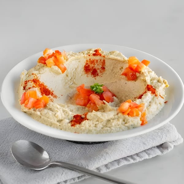 Creamy hummus in a white bowl, garnished with diced tomatoes, paprika, and parsley, with a spoon.