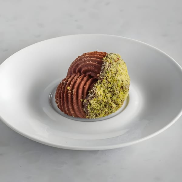 A chocolate dessert, half covered in green pistachio crumbles, on a white plate.