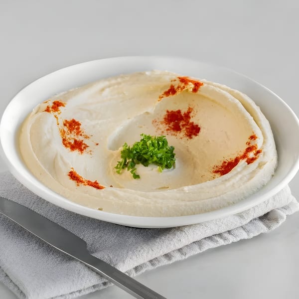 Creamy hummus in a white bowl, garnished with paprika and fresh parsley, on a gray napkin.